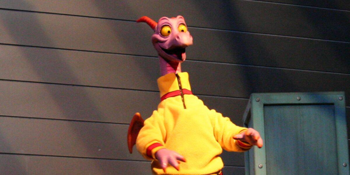 A purple cartoon dragon with orange wings, wearing a yellow sweater, stands in front of a gray paneled wall and a blue prop box, smiling with wide yellow eyes in true Disney style.