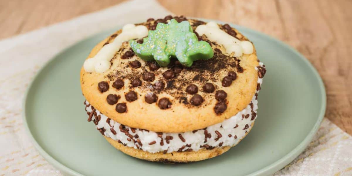 A Disney-inspired dessert sandwich with chocolate chip cookies, rolled in chocolate sprinkles and filled with cream, topped with icing dog bone shapes and a green icing decoration on a light green plate.