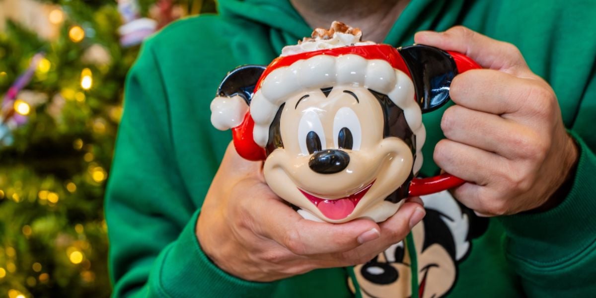A person wearing a green hoodie with a cartoon character design holds a festive holiday mug shaped like a smiling Mickey Mouse with a Santa hat, in front of a blurred Christmas tree.