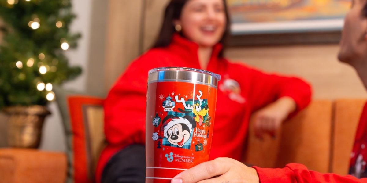 A hand holds a red Disney-themed holiday tumbler featuring Mickey Mouse and festive designs. In the background, two people in red sweaters sit on an orange couch, smiling next to a decorated Christmas tree.