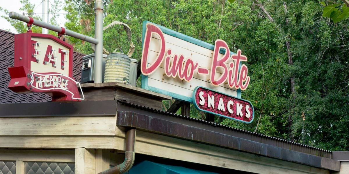 A vintage-style sign reads “Dino-Bite Snacks” above a Disney snack stand, with a red “EAT” sign and lush green trees in the background. The building features a rustic, wooden exterior.