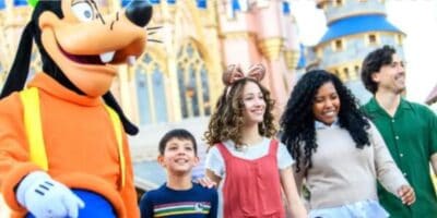 A group of four people, including two children and two adults, walk happily with a person in a Goofy costume at a theme park, with a fairytale castle visible in the background.