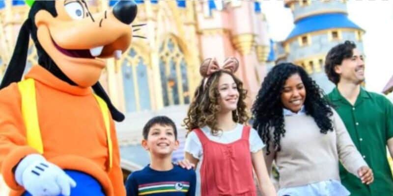A group of four people, including two children and two adults, walk happily with a person in a Goofy costume at a theme park, with a fairytale castle visible in the background.
