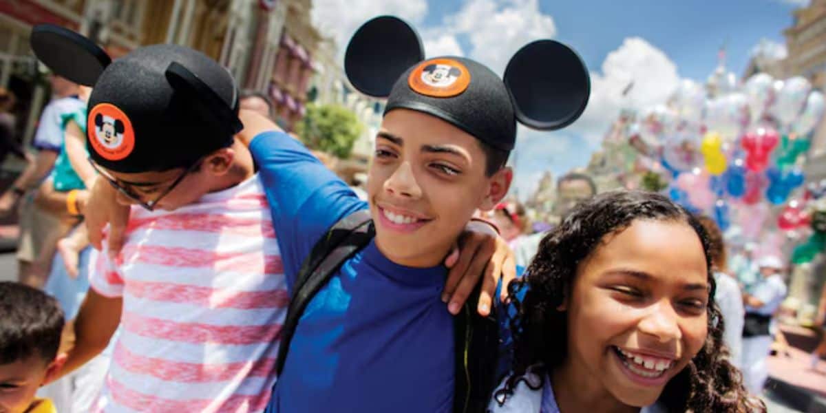Three smiling kids walk arm-in-arm at a Disney theme park, two wearing Mickey Mouse ear hats. Colorful balloons and blue sky are visible in the background.