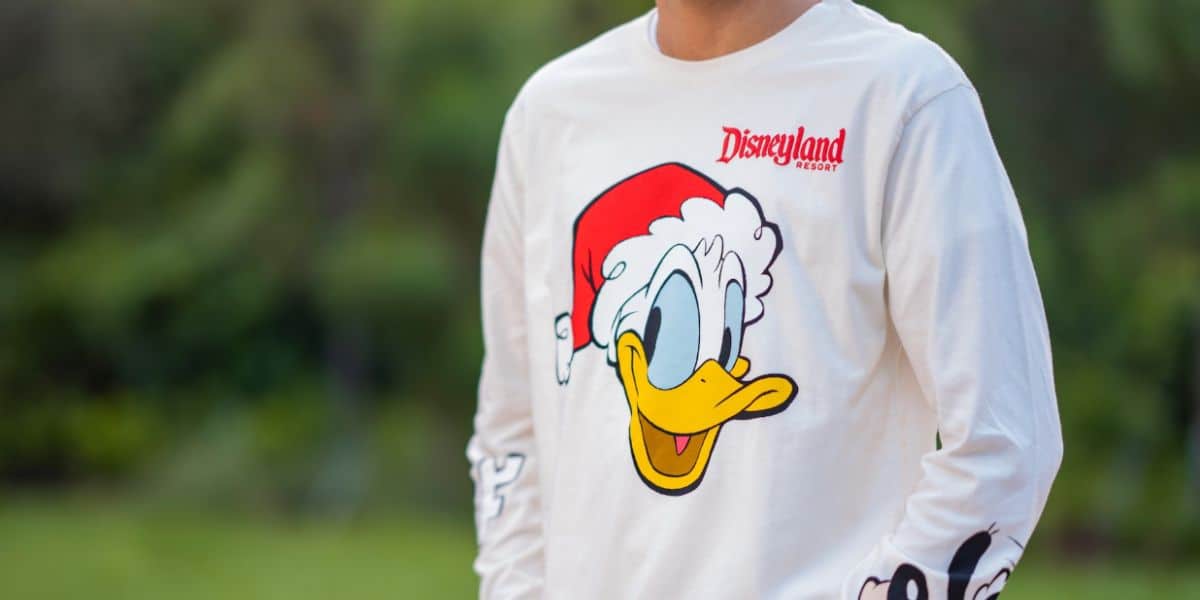 A person wearing a white shirt featuring a large image of Donald Duck in a Santa hat and the red "Disneyland Resort" logo stands outdoors, capturing the fun and festive holiday spirit against a blurred green background.