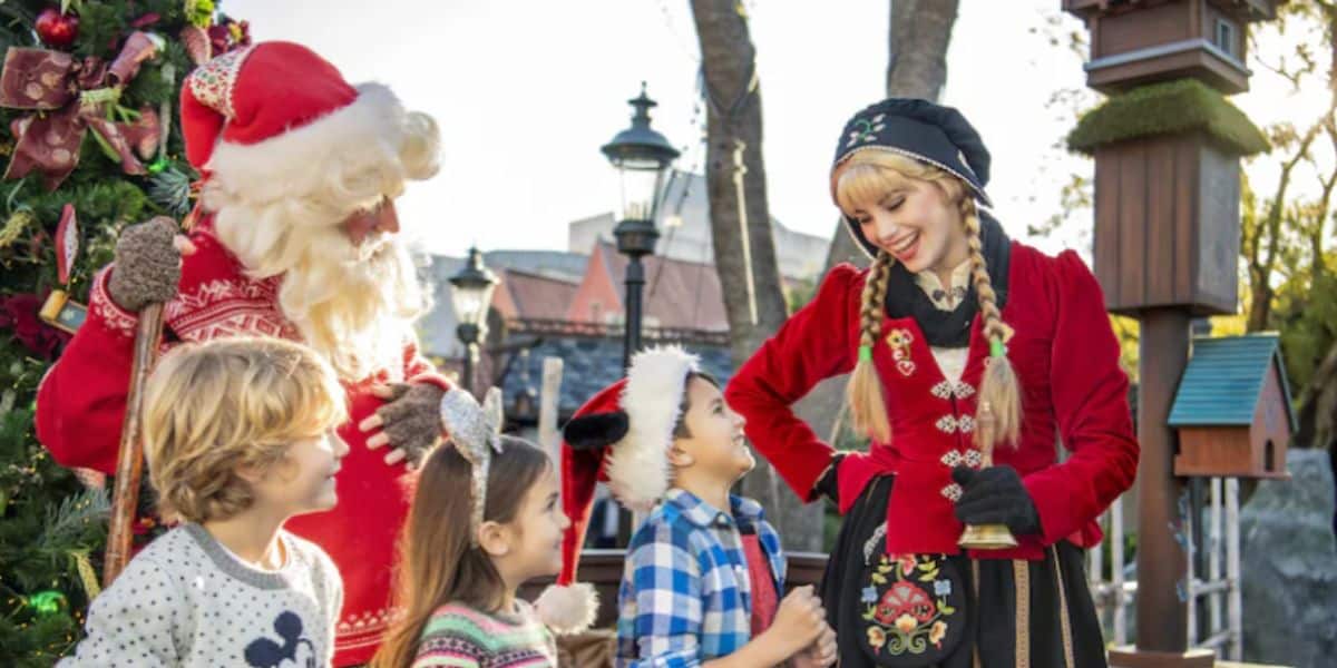 Santa Claus and a woman in festive costume stand beside a Christmas tree at EPCOT, smiling and interacting with three children wearing holiday attire, including Santa hats and reindeer ears, in an outdoor setting.