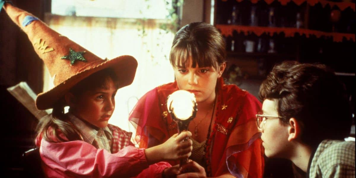 Three children in costumes, one wearing a pointed witch hat, sit around a glowing orb, gazing intently at it in a Halloween-themed room filled with Disney-inspired decorations.