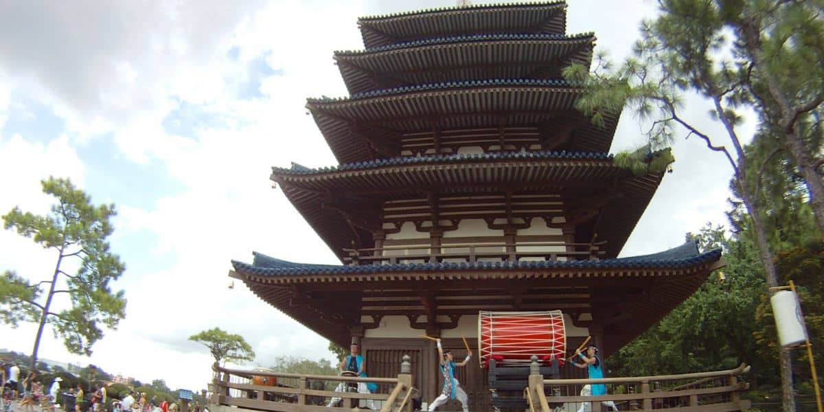 A large wooden pagoda stands tall with multiple tiers. In front, a group of people in blue outfits play a large red drum on a raised platform. Green trees surround the scene under a partly cloudy sky.