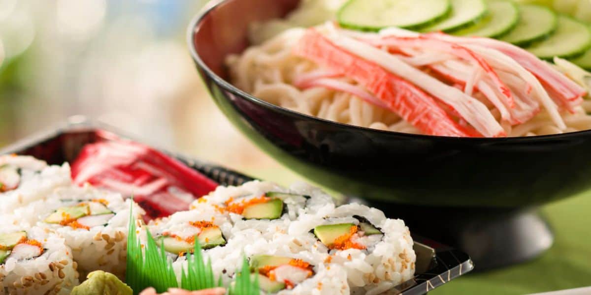 A close-up of sushi rolls filled with crab, cucumber, and rice, with a side of wasabi and pickled ginger. In the background, a bowl features Japan-inspired ingredients like sliced cucumber, imitation crab sticks, and noodles.