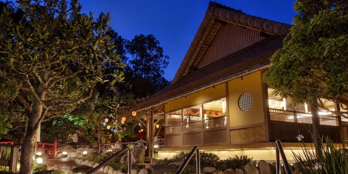 A warmly lit traditional Japanese-style building with a thatched roof is surrounded by trees and hanging lanterns at dusk, capturing the serene and inviting spirit of Japan.