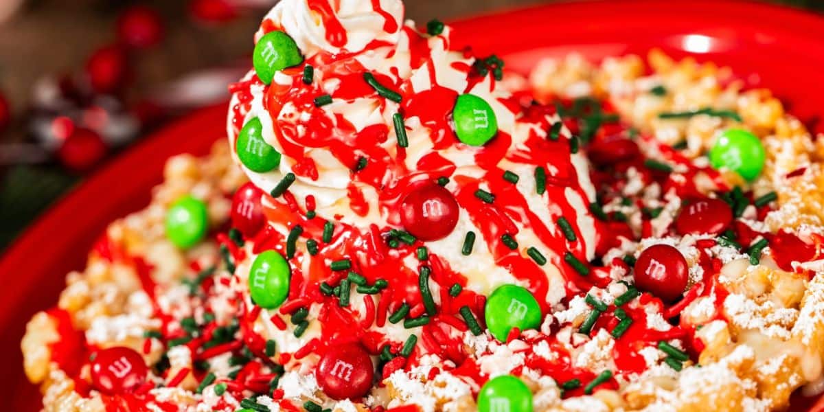 A close-up of festive dessert popcorn topped with whipped cream, red and green M&Ms, red syrup, green sprinkles, and powdered sugar, served on a red plate—perfectly capturing the magic of a Disney World holiday treat.