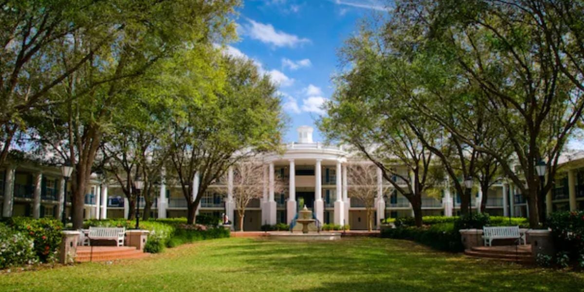 A large building with white columns and a circular entrance, reminiscent of grand hotels, is framed by tall trees and a grassy courtyard, with benches and a central fountain under a blue sky with clouds.