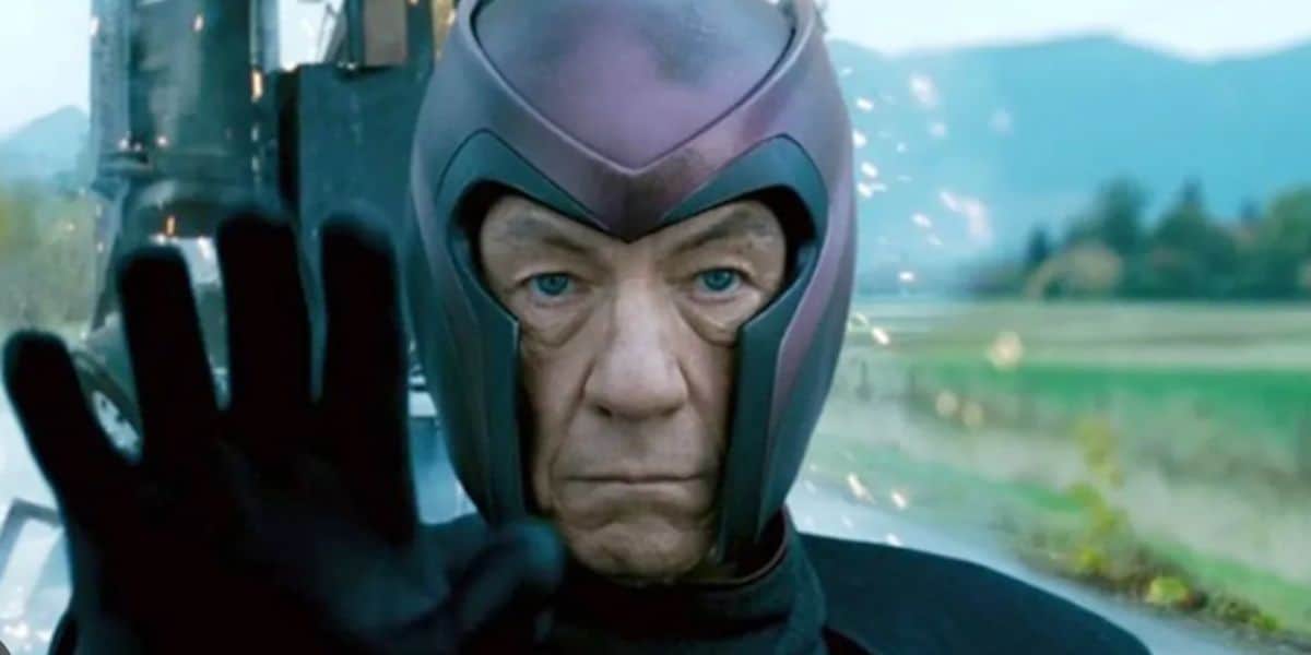 An older man, resembling Robert Downey Jr., in a metallic helmet and dark suit raises his hand with a serious expression as sparks fly behind him. He stands outdoors, with distant mountains and greenery in the background.