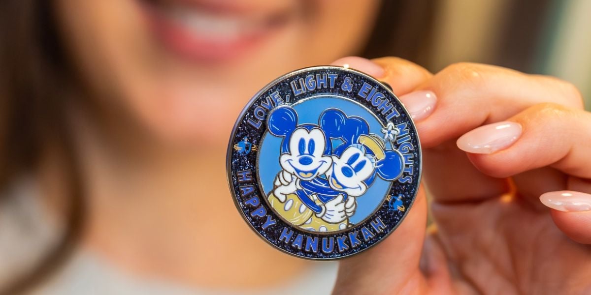 A person holds a round holiday pin featuring Mickey and Minnie Mouse in blue and white, with the text "Love, Light & Eight Nights. Happy Hanukkah.
