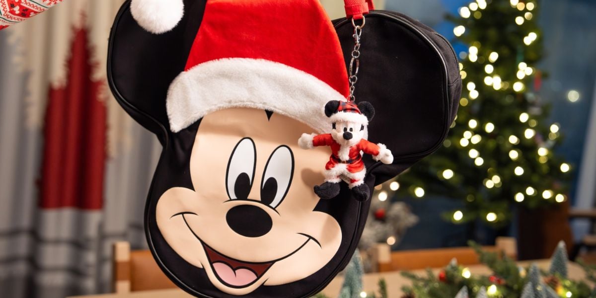 A holiday-themed Mickey Mouse-shaped bag with a Santa hat and a small plush Mickey Mouse in a Santa outfit hanging from it, set in a festive room with a decorated Christmas tree in the background.