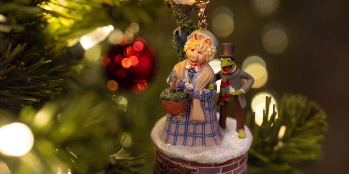 A close-up of a holiday Christmas tree ornament featuring two characters, including a green frog in a top hat and a woman in a bonnet, hanging on a decorated tree with blurred lights and a red bauble in the background.