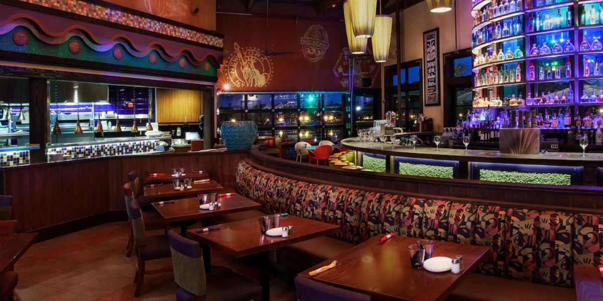 A stylish restaurant interior with wooden tables, patterned booth seating, a curved bar lined with colorful backlit shelves of bottles, and large pendant lights, creating a vibrant and modern restaurant atmosphere.