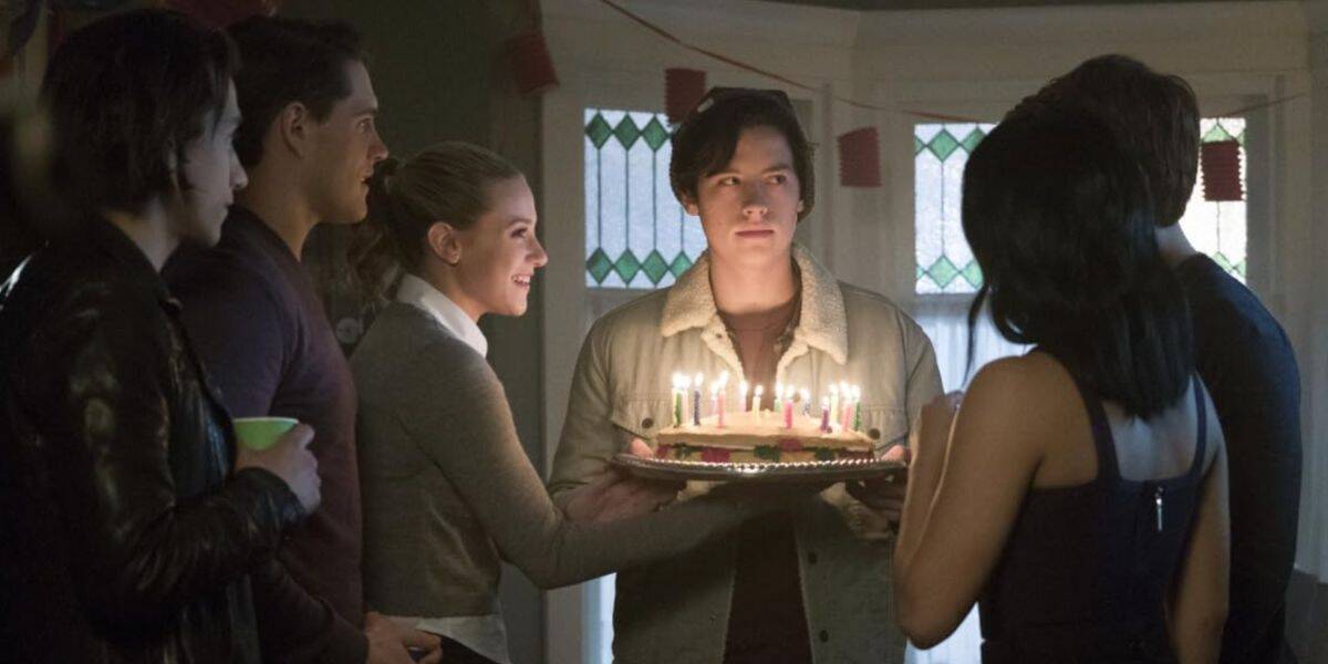 A group of young adults gather indoors, Riverdale style, around a person holding a cake with lit candles, celebrating a birthday. The softly lit room features stained glass windows and red decorations in the background.
