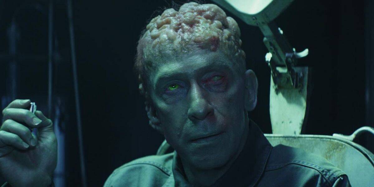 A man with a deformed, exposed, swollen brain and glowing green and red eyes sits in a chair, holding a cigarette, under eerie, dim lighting—his tattered Captain America shirt hinting at a lost sense of heroism.