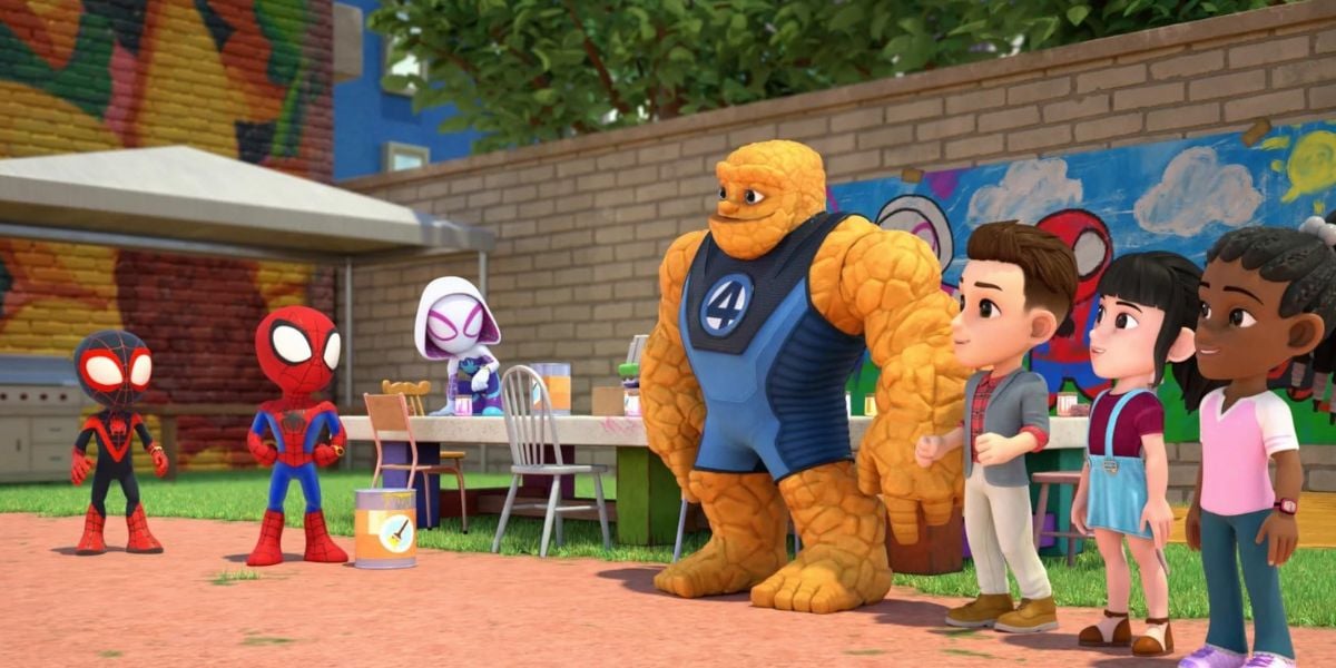 Animated superheroes, including Spider-Man, Miles Morales, and the Thing, stand outside near children by a painted wall and tables with art supplies, interacting with kids at a colorful outdoor Spider-Man-themed event.