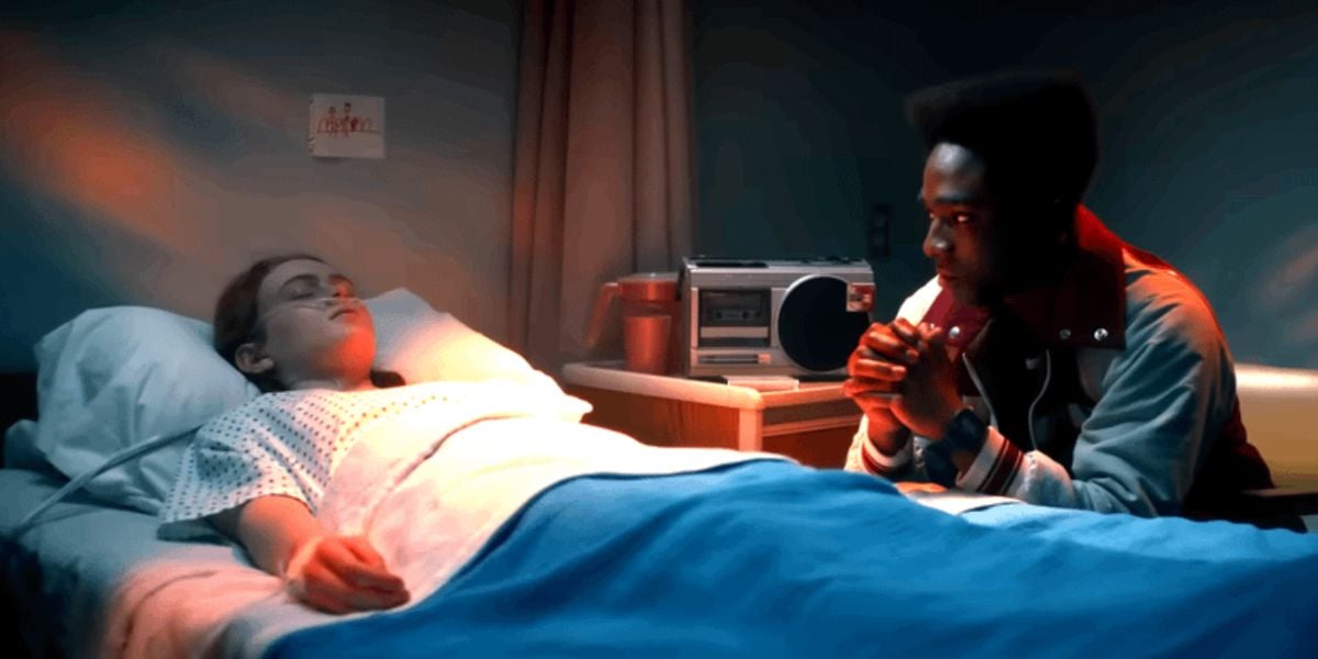 A young girl lies unconscious in a hospital bed under a blue blanket, while a concerned young man, reminiscent of Stranger Things, sits beside her, hands clasped and watching her intently in the dimly lit room.