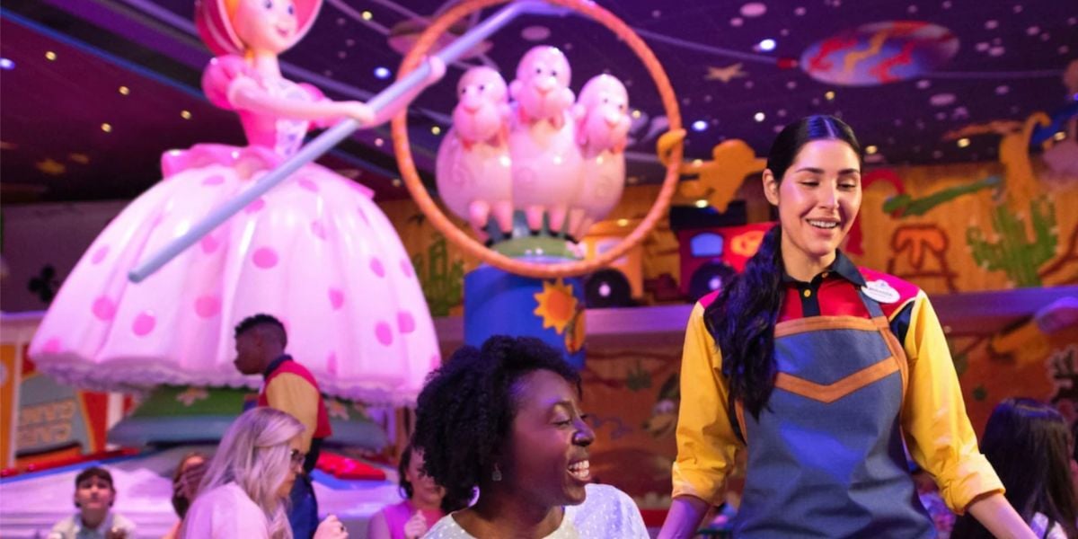 A theme park worker in a colorful uniform smiles while assisting a guest near a large statue of Bo Peep with sheep. Other visitors enjoy the vibrant, Toy Story-themed attraction and nearby restaurant offerings.