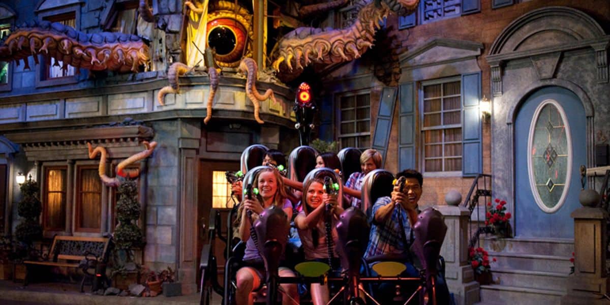 A group of people ride an indoor amusement park attraction, aiming laser guns at targets, with a large, monstrous tentacled creature—reminiscent of Men in Black—displayed above them as part of the themed decor.