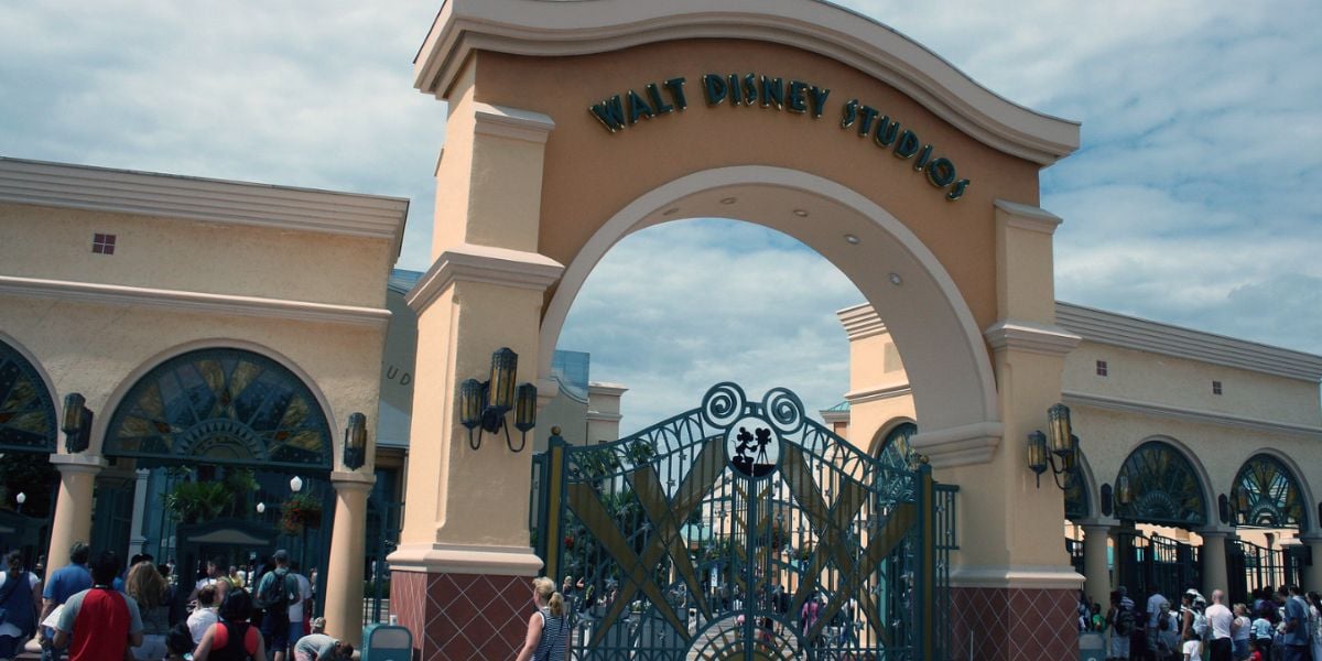 Large entrance gate to Walt Disney Studios with golden lettering above an arched gateway, ornate ironwork, and people walking underneath on a partly cloudy day at Walt Disney Studios.