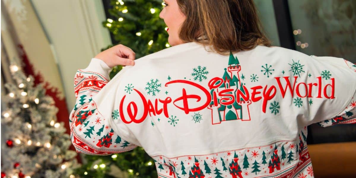 A person wearing a festive holiday sweater with "Walt Disney World" and a castle design on the back stands near a decorated Christmas tree inside a cozy room.