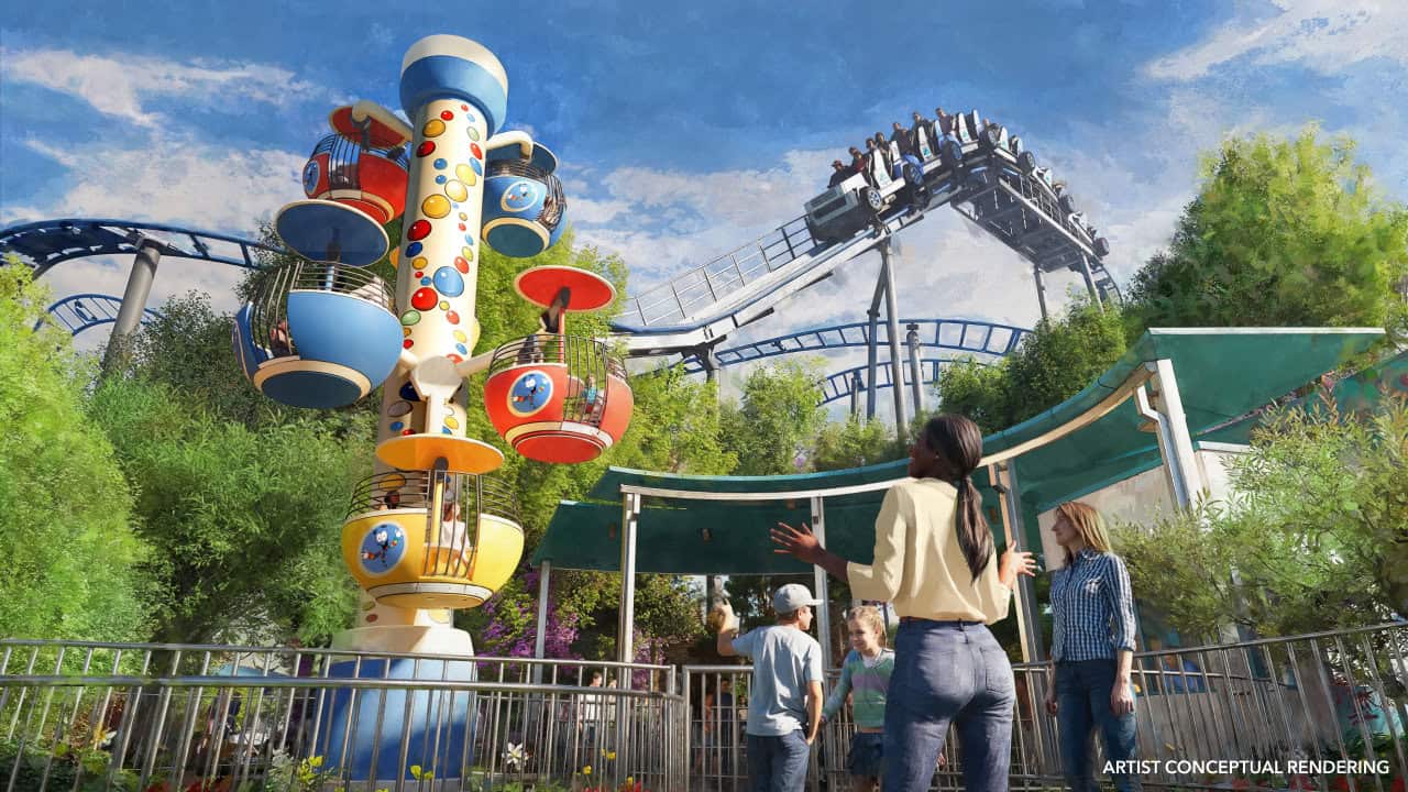 Artist rendering of a theme park with a colorful spinning tower ride in the foreground, several people admiring it, and a roller coaster with riders in the background, surrounded by greenery, showing Jurassic World inside of a new Universal theme park coming soon.