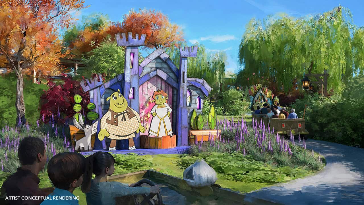A colorful theme park ride passes by a whimsical gingerbread house facade featuring cartoon ogre characters, surrounded by lush trees, purple flowers, and vibrant landscaping under a sunny sky showing Shrek and characters inside of Universal Kids Resort.