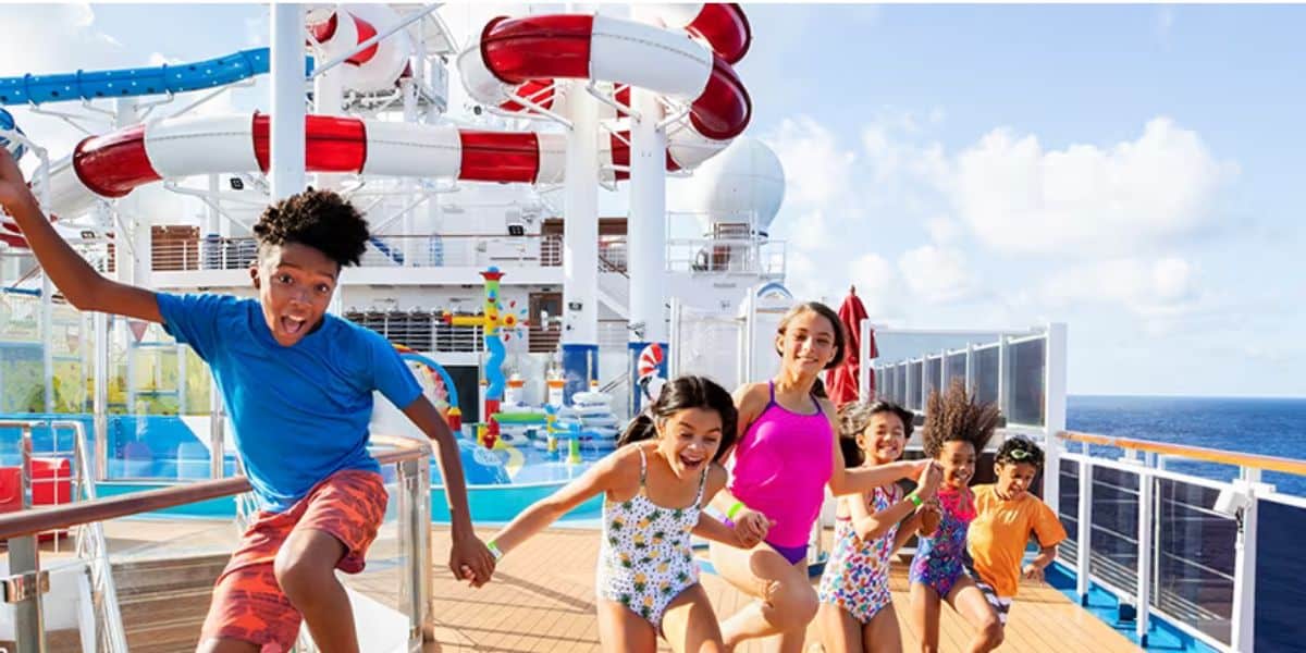 Six children in swimsuits run and smile on a cruise ship deck near a colorful water park and large water slide, with the ocean and blue sky in the background.