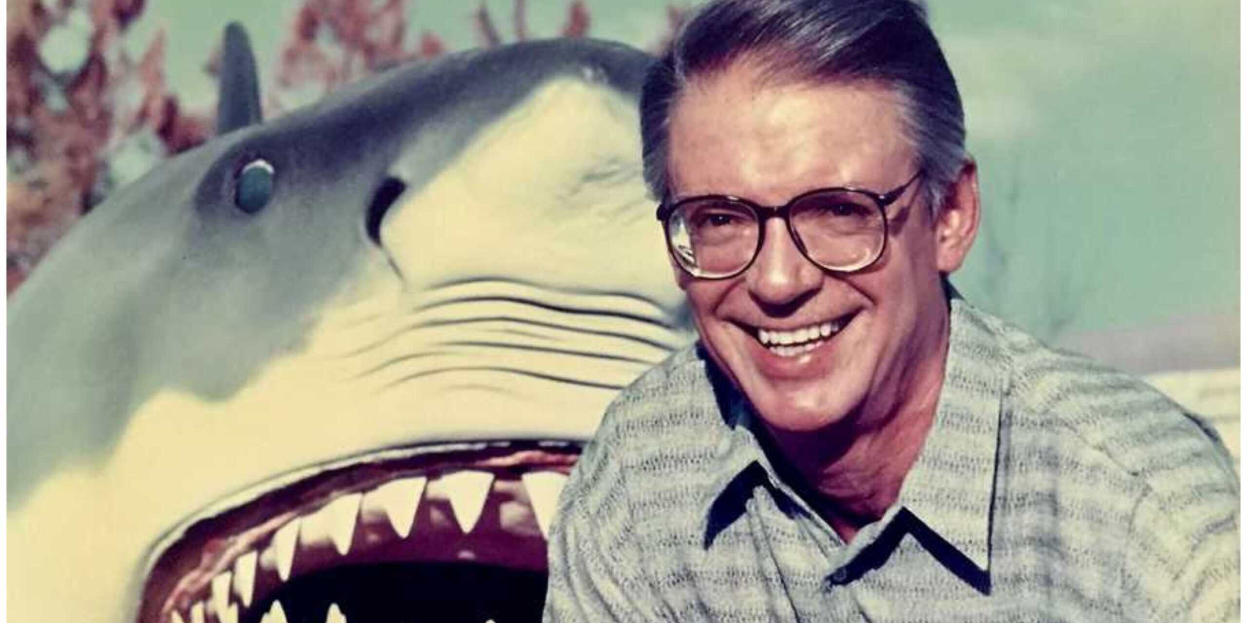 A smiling man with glasses stands in front of a large, open-mouthed shark model or statue outdoors, with trees and a blue sky in the background.