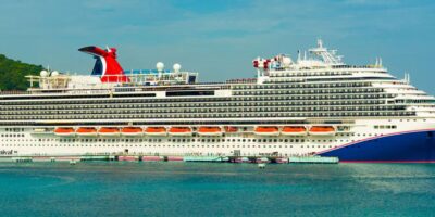 A large white cruise ship with multiple decks and red funnel docked at a port, with calm blue water in the foreground and a green hillside in the background.