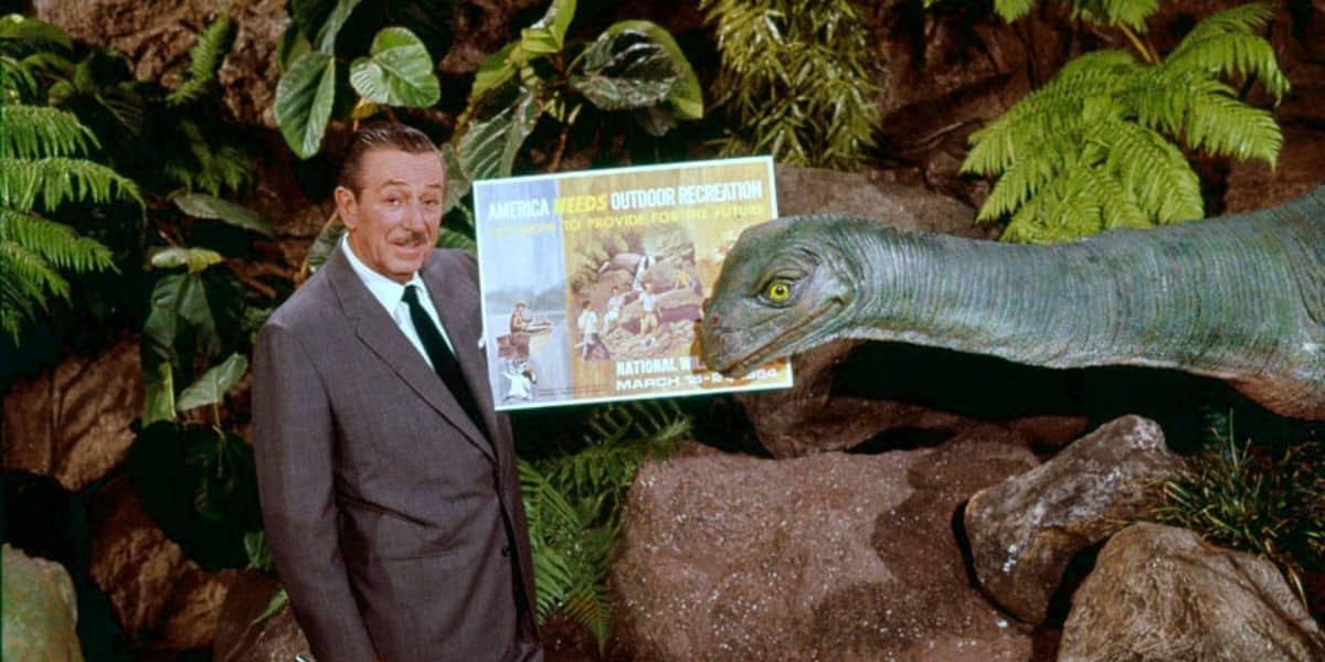 A park host in a suit welcomes guests beside a lifelike dinosaur exhibit and vibrant poster in a jungle-themed attraction.