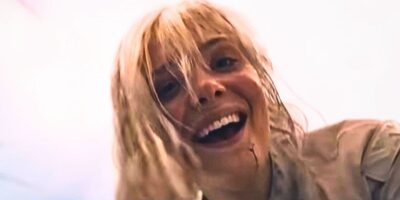A woman with messy blonde hair smiles widely at the camera; her face and teeth are visible up close, with a small streak of blood near her mouth, evoking the raw tension of Predator: Badlands. The background is bright and out of focus.