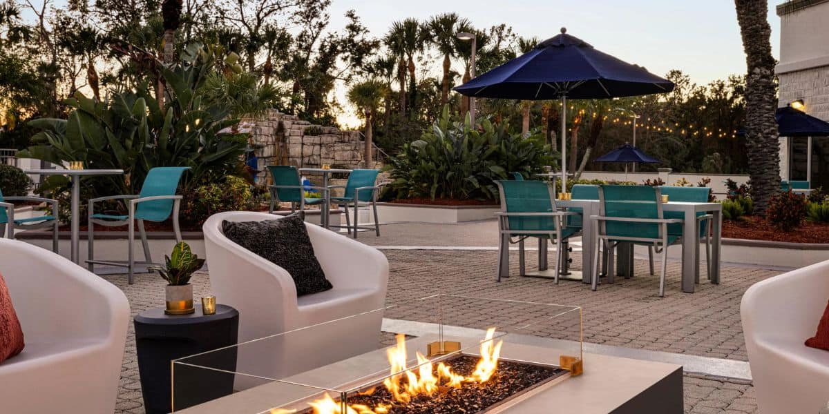 Outdoor patio with modern fire pit, white and teal chairs, blue umbrellas, small tables, and lush landscaping. String lights hang above, and trees line the background at sunset. Cozy, inviting atmosphere.
