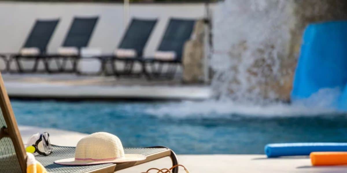 A sunhat and sunglasses rest on a lounge chair beside a swimming pool, with water splashing from a feature and pool noodles floating nearby. Several empty lounge chairs are visible in the background.