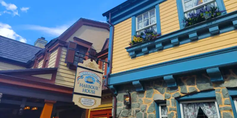 A sign reading "Columbia Harbour House" hangs between two charming colonial-style buildings with yellow siding, blue trim, stone accents, and flower boxes under windows. The sky above is blue with scattered clouds.