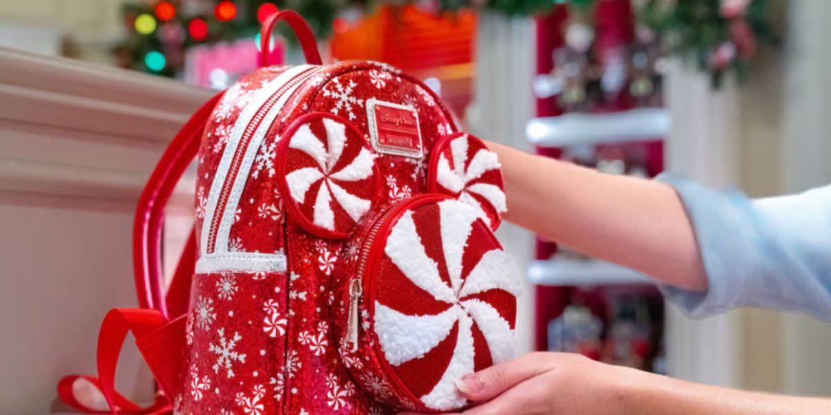 A person holds a festive red Disney backpack decorated with white snowflakes and peppermint swirl designs, featuring mouse ear-shaped compartments, in a brightly lit holiday-themed store.