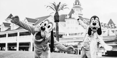 Two people in Pluto and Goofy costumes pose with arms outstretched in front of a Victorian-style riverside building with a tower and decorative roof, possibly at a theme park. The black and white image evokes a nostalgic charm.