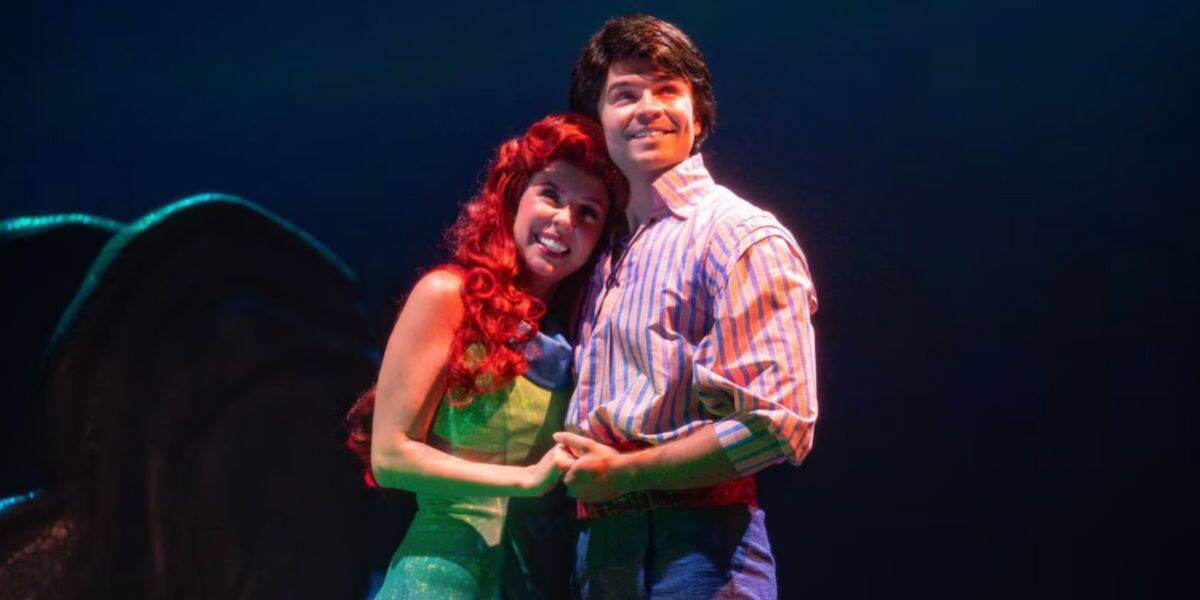 Two actors on stage, dressed as Ariel and Prince Eric from Disney’s "The Little Mermaid," stand close together, smiling and holding hands, with blue lighting creating a magical atmosphere behind them.
