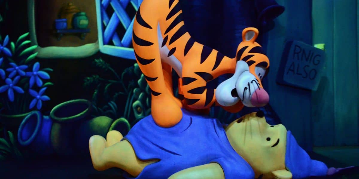 Tigger playfully bounces on top of Winnie the Pooh, who is lying on the ground in front of a house surrounded by colorful plants and pots. A "Ring Also" sign is visible on the right, adding charm to this Winnie the Pooh scene.
