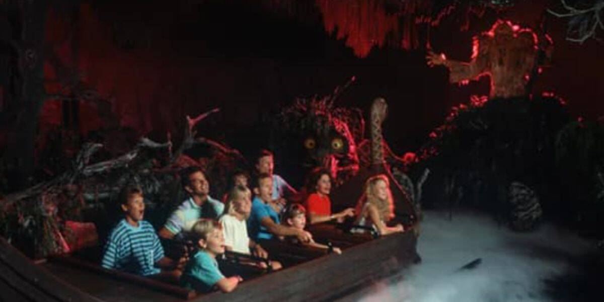 A group of people ride a log flume attraction, looking surprised as they approach a maelstrom-lit, illuminated tree figure in a dark, misty, and spooky setting.