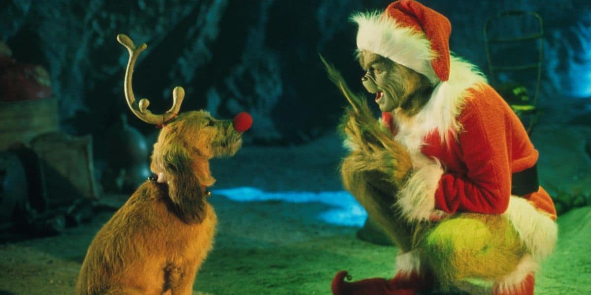 A furry green Grinch dressed as Santa Claus excitedly faces a brown dog wearing antlers and a red nose in a cave-like setting with green lighting.