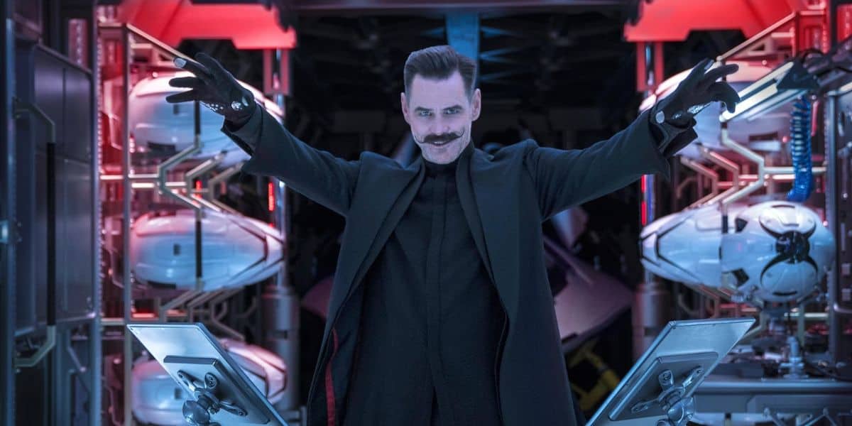 A man with a dark coat and gloves stands with his arms raised in a high-tech lab, surrounded by futuristic robotic devices and illuminated by red and blue lights. With slicked-back hair, a handlebar mustache, and a sly grin, he exudes a grinch-like charm.