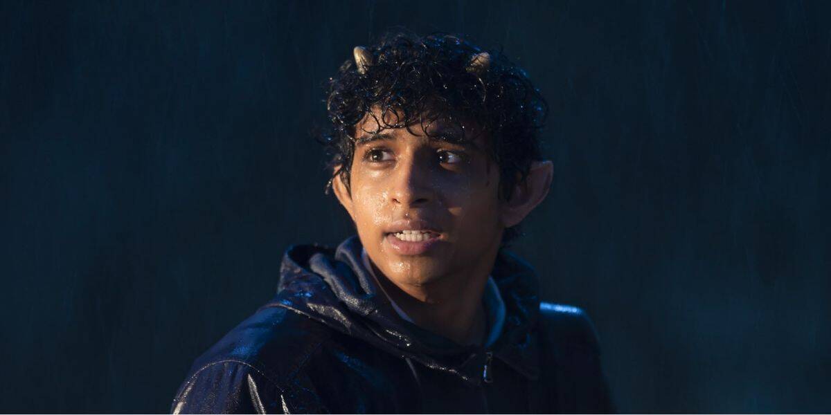 A young person with small horns on their head and pointed ears stands in the rain, looking concerned—reminiscent of a Percy Jackson character. Their wet, curly hair clings to their face, and they wear a dark, hooded jacket.