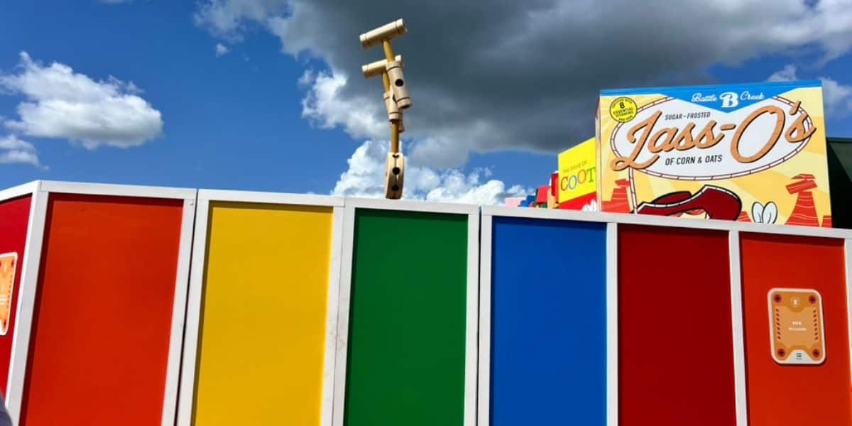 A colorful wall with vertical panels in red, yellow, green, and blue sits under a partly cloudy sky, evoking a playful Disney vibe. Behind the wall is a large fake cereal box labeled "Lasi O’s" and a wooden toy periscope.
