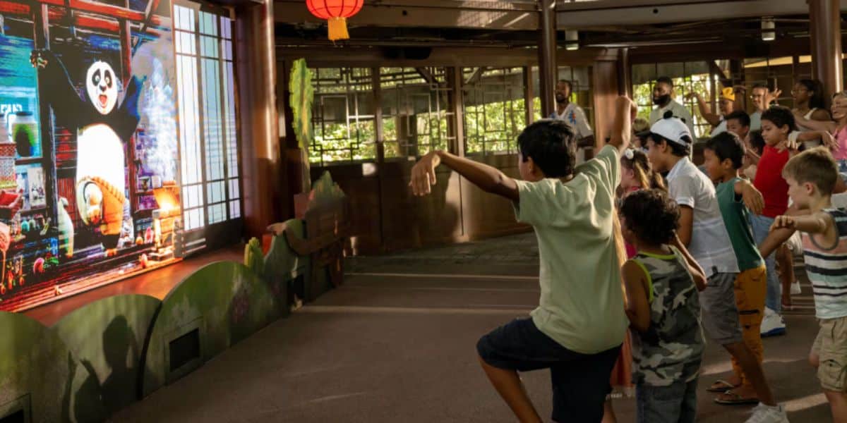 A group of children stand indoors mimicking martial arts poses while watching a DreamWorks animated panda character on a large screen, with adults observing in the background.
