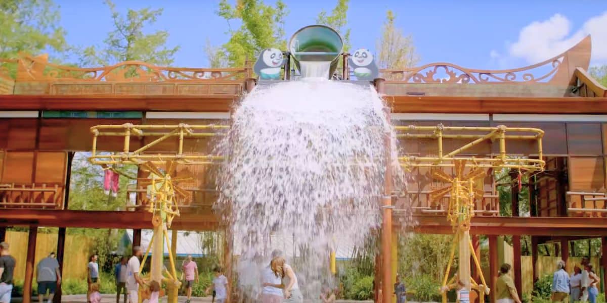 A large bucket of water tips over, splashing down from a DreamWorks-themed water play structure adorned with smiling panda faces. People, including children, stand below enjoying the playful splash on a sunny day.